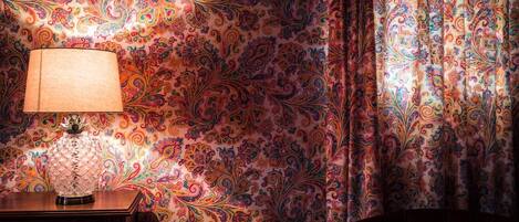 Une pièce avec un papier peint à motifs, une lampe posée sur une table d’appoint en bois et un rideau orné de motifs cachemire.