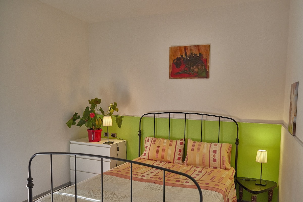 Ein Zimmer mit einem Bett, einem Nachttisch, einer Lampe, einem Topfpflanze und einem Bild an der Wand.