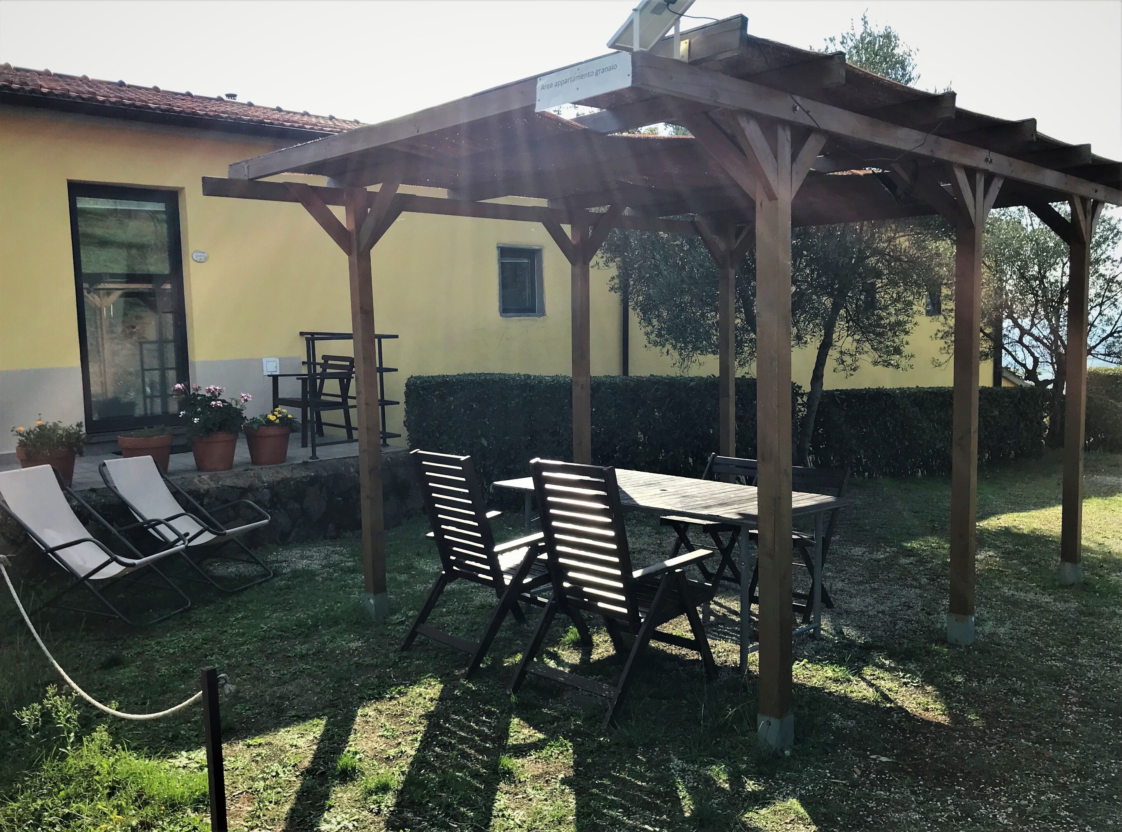 Eine überdachte Terrasse mit Holztisch und -stühlen, Blumentöpfen und einem gelben Gebäude im Hintergrund.