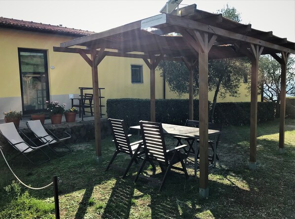 Un patio cubierto con una mesa y sillas de madera, plantas en macetas y un edificio amarillo al fondo.