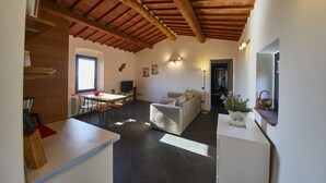 A modern living area with a sofa, dining table, and chairs. There is a kitchen area with a countertop and a sink. The ceiling has exposed wooden beams. - Colle Da Vinci (Vinci)