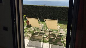 A view through an open door to a patio with two beige lounge chairs, a hedge, and a distant view of the sea. - Colle Da Vinci (Vinci)