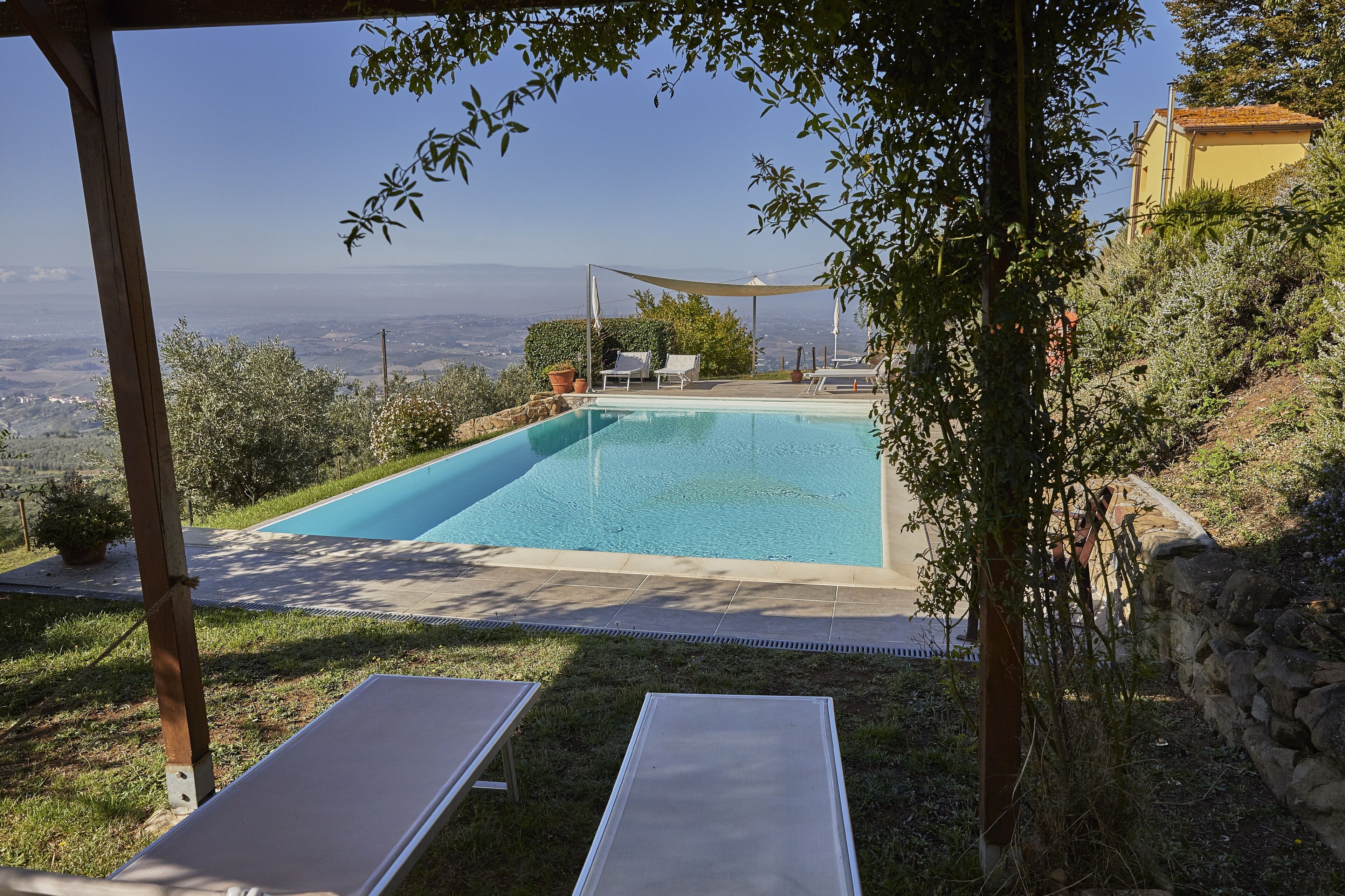 A swimming pool with a pergola and lounge chairs, surrounded by greenery and overlooking a distant landscape.