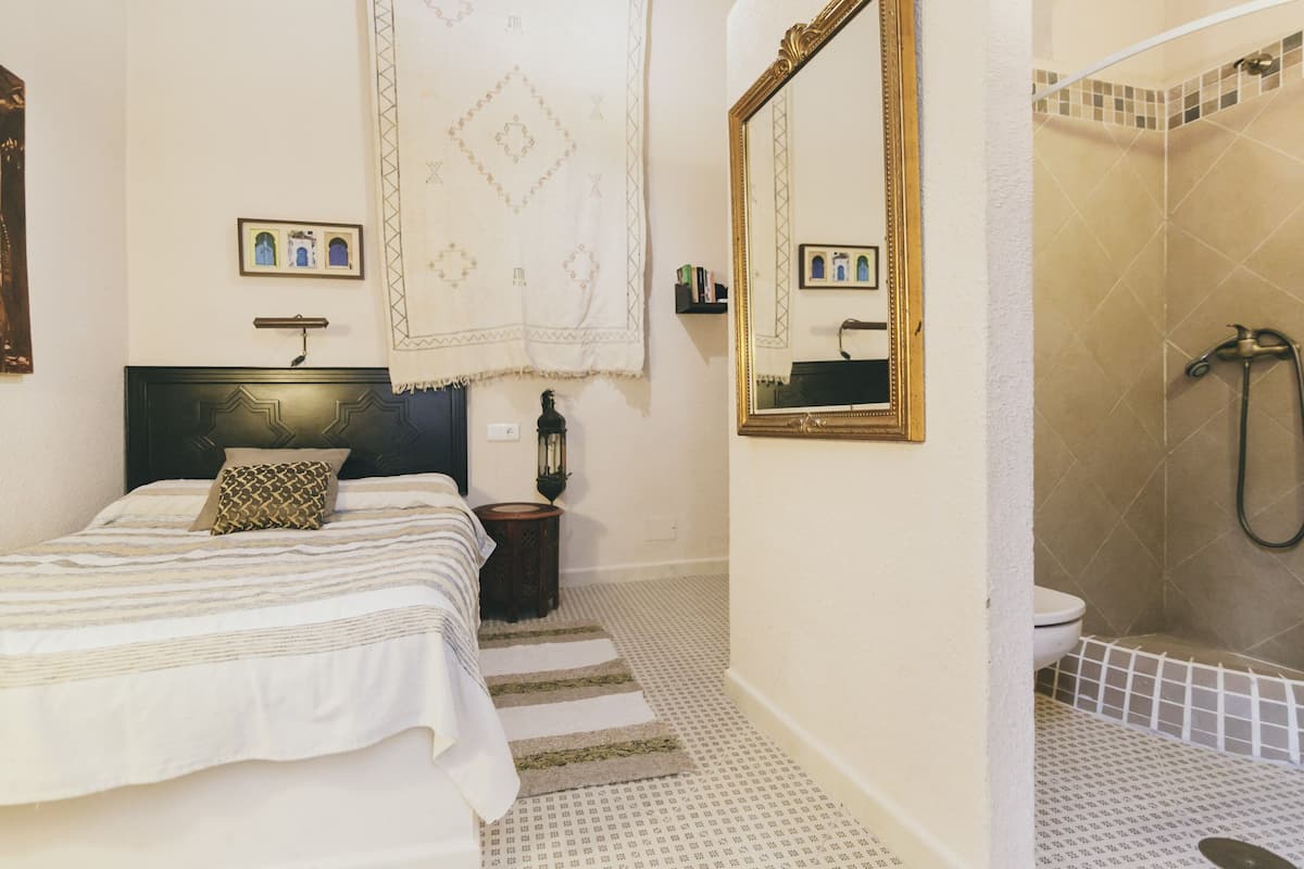 A bedroom with a bed, a nightstand, a mirror, and a bathroom with a shower and toilet.