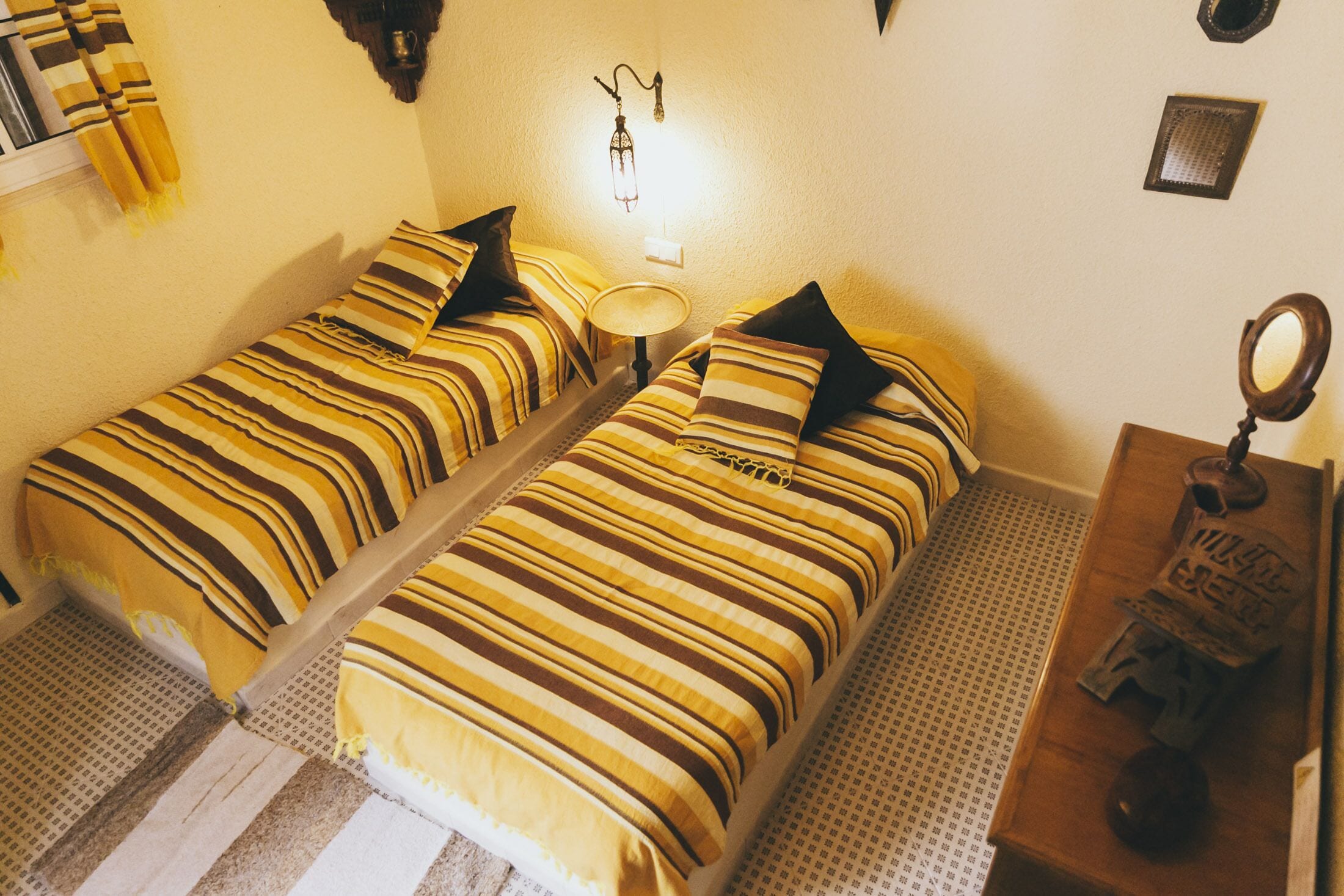 A room with two beds, each with striped bedding and black pillows. There is a small round table with a lamp between the beds, and a wooden dresser with a mirror and a small object on top.
