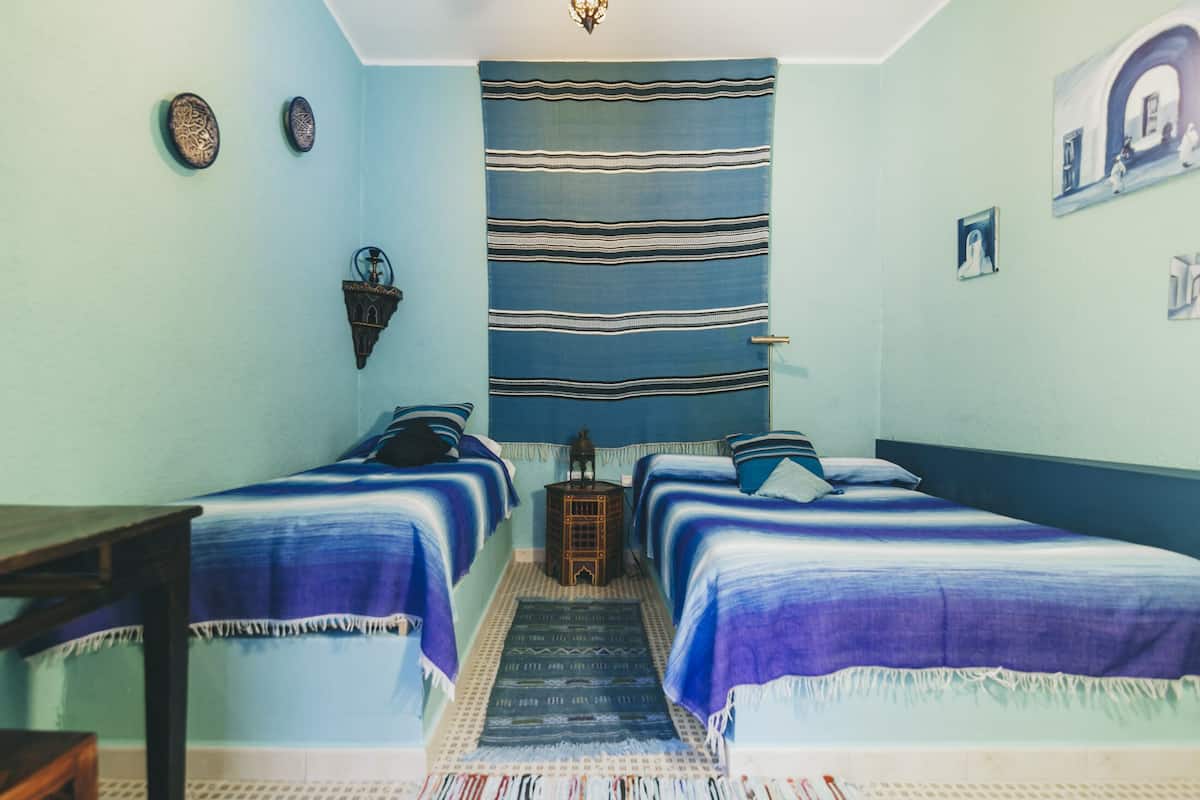A room with two beds, a small table, a wall-mounted lamp, and a patterned rug.