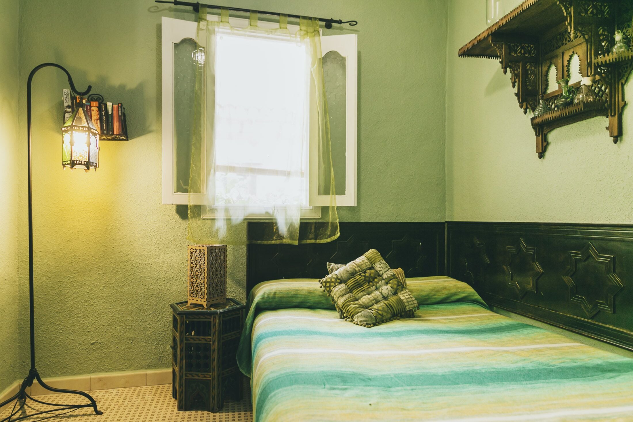 A room with a bed, a nightstand, a floor lamp, a window with curtains, and a wall-mounted shelf.
