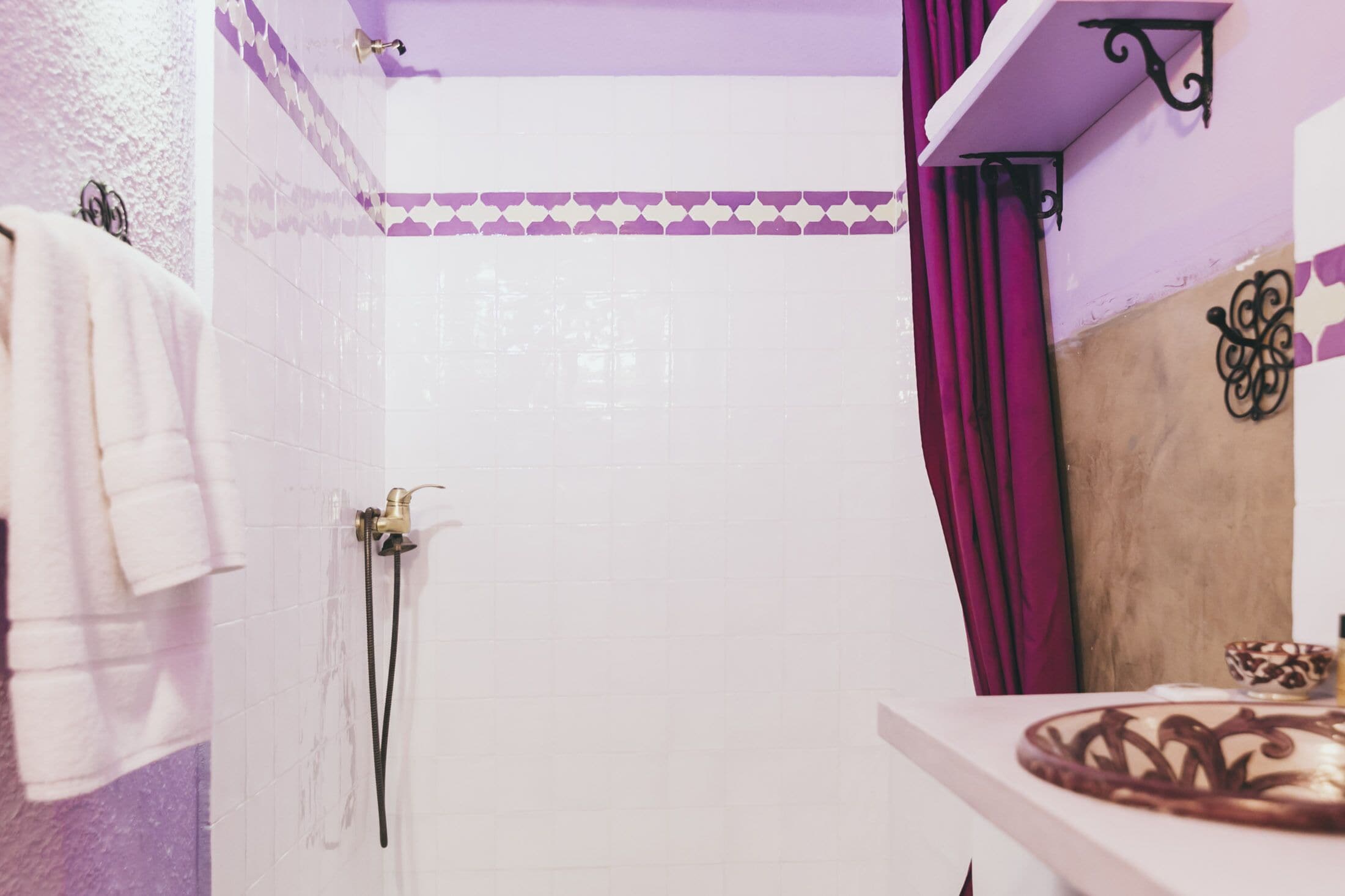 Une salle de bain avec des murs recouverts de carreaux blancs, un espace douche, un meuble-lavabo agrémenté d’un vase décoratif et un rideau violet.