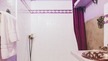 Une salle de bain avec des murs recouverts de carreaux blancs, un espace douche, un meuble-lavabo agrémenté d’un vase décoratif et un rideau violet.