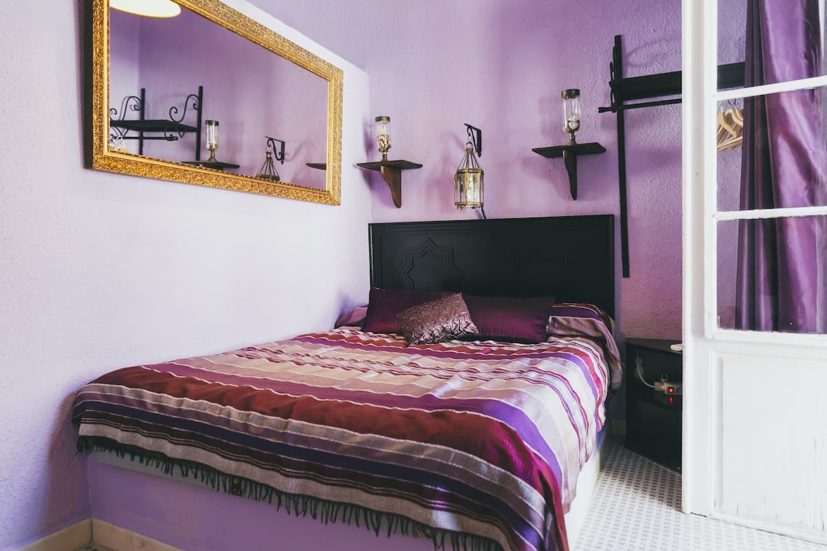 A bedroom with a bed, a large mirror, wall-mounted sconces, and a window with curtains.