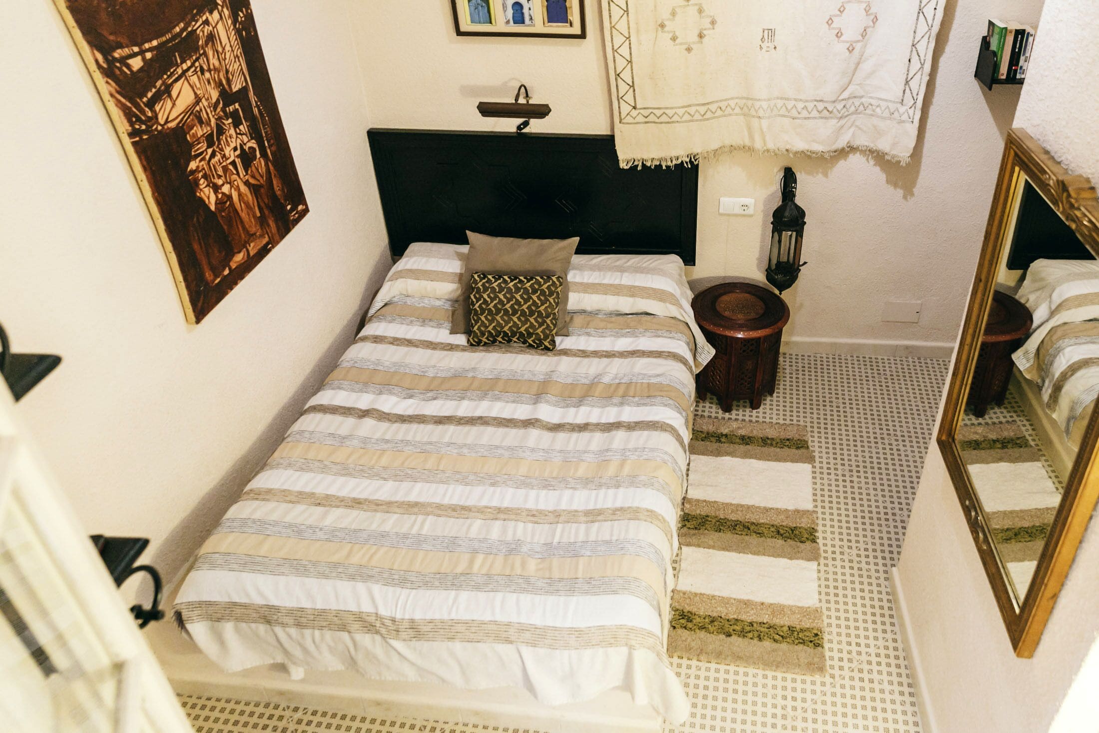 Un lit bien fait, avec une literie à rayures, une tête de lit, une petite table de chevet avec une lampe, un coussin à motifs, un tapis, un tableau accroché au mur et un miroir.