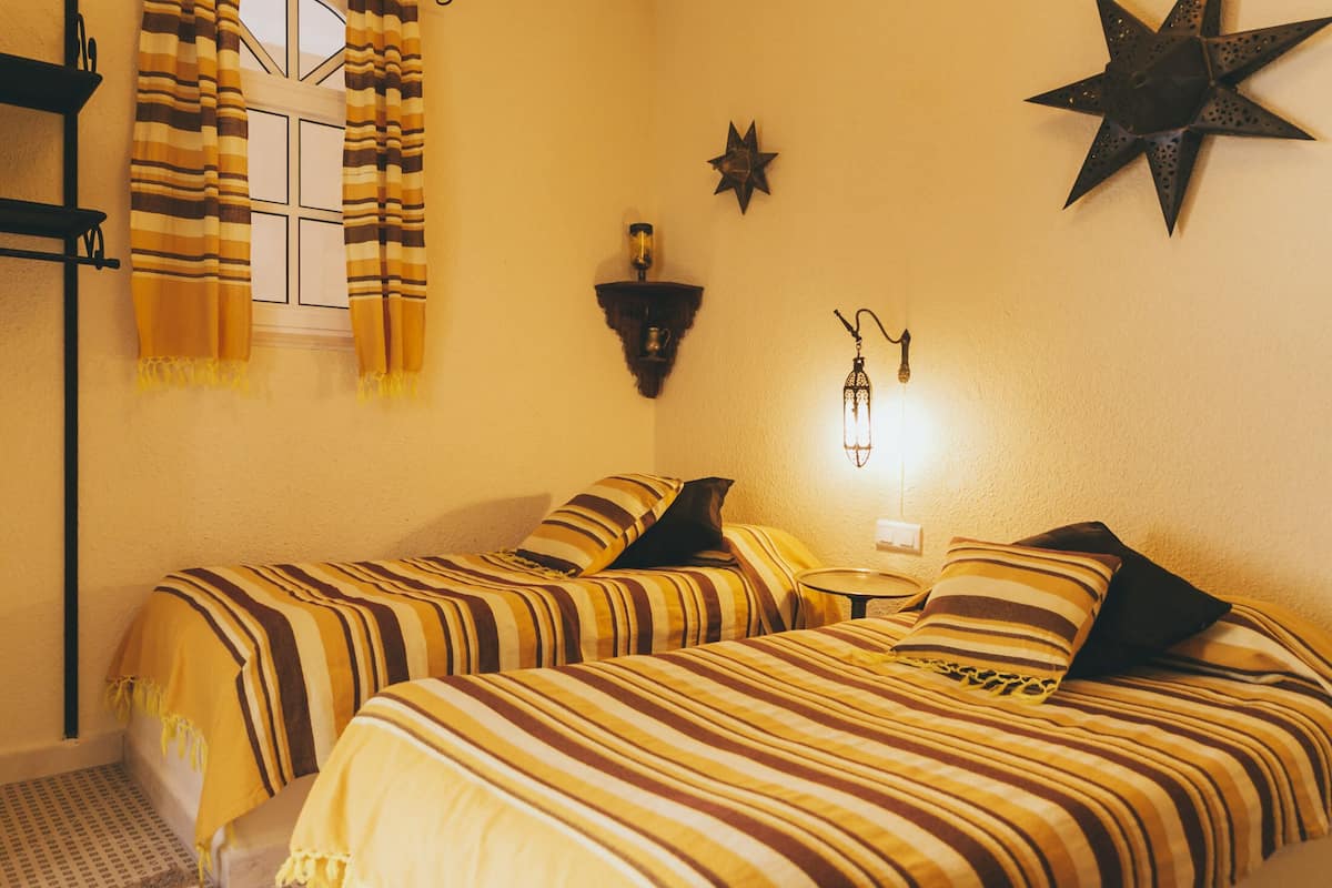 A room with two beds, striped bedding, a star-shaped wall decoration, a wall-mounted light fixture, and a window with curtains.