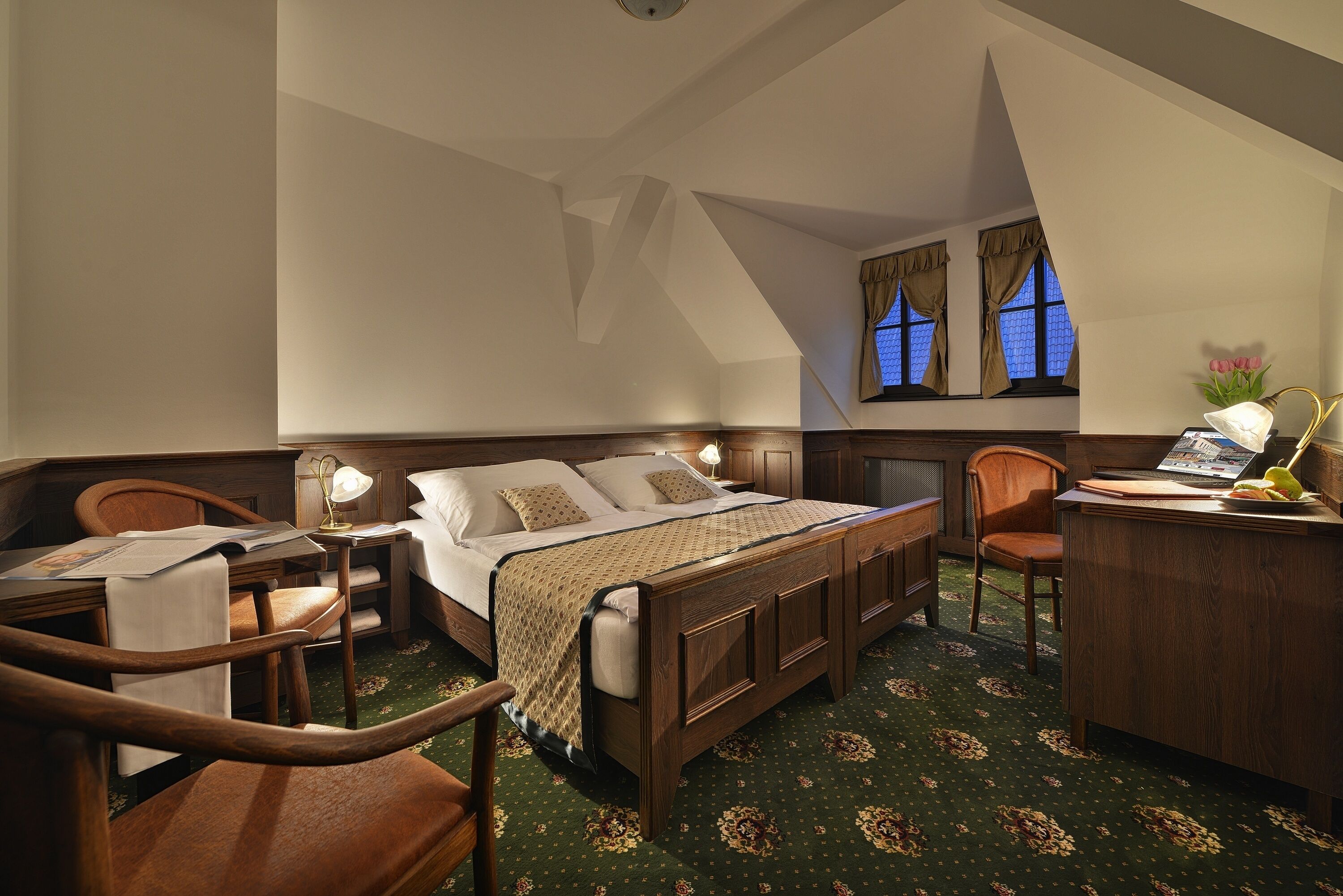 A hotel room with a wooden bed, a desk with a lamp, a chair, and a window with curtains.