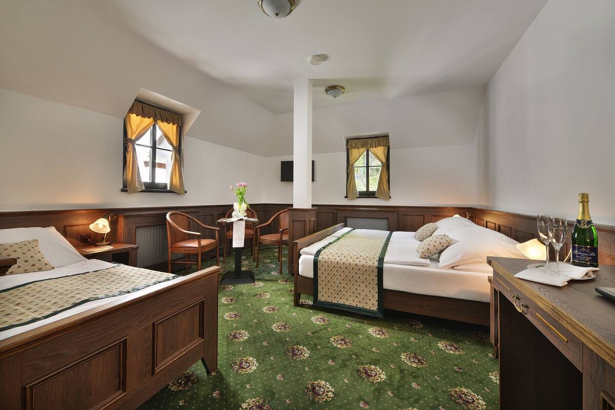 Ein Hotelzimmer mit zwei Betten, einem Schreibtisch, einem Sessel, einem kleinen Tischchen mit einer Vase und einer Champagnerflasche auf dem Schreibtisch.