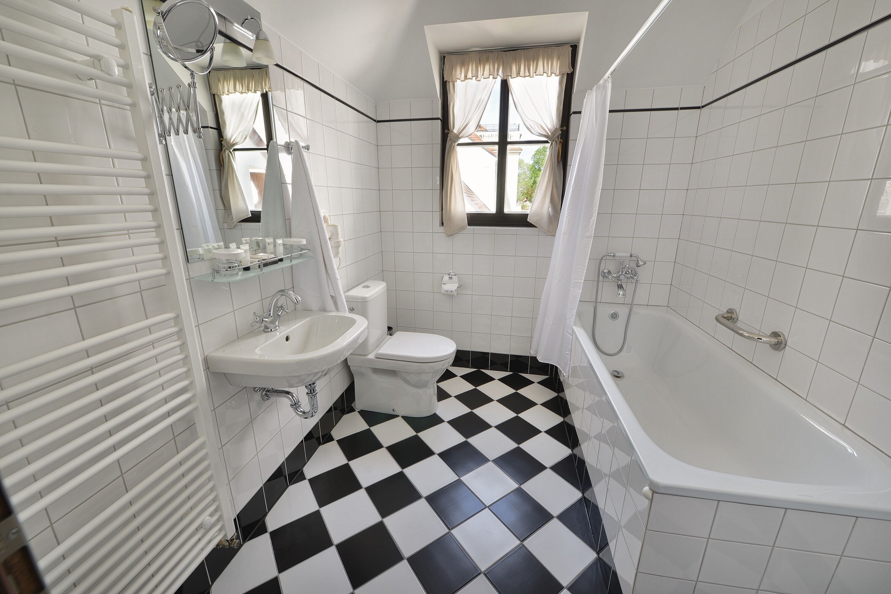 A bathroom with a checkered floor, white tiles, a bathtub, a toilet, a sink, and a window with curtains.
