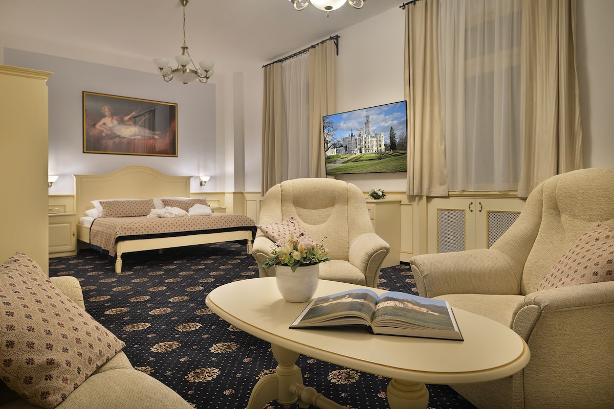 Ein Hotelzimmer mit einem Bett, zwei Sesseln, einem kleinen Tisch mit einem Buch und Blumen, einem Bild an der Wand und einem Fernseher.