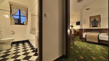Ein Hotelzimmer mit Schachbrettmuster auf dem Boden, ein Badezimmer mit Toilette und Waschbecken, ein Bett mit einem gemusterten Überwurf und ein kleiner Tisch mit einer Blumenvase.