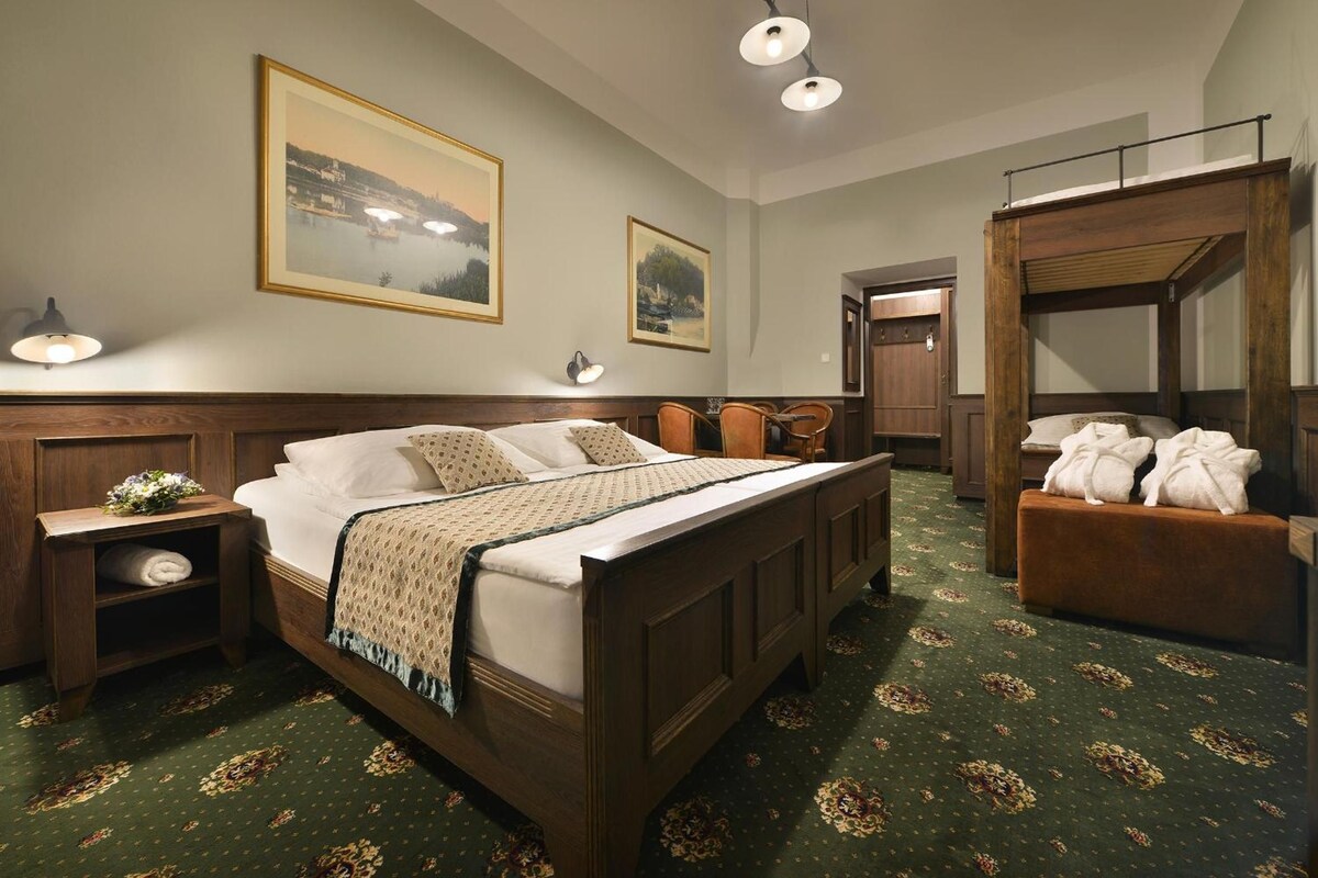 Une chambre d’hôtel avec un grand lit, une tête de lit en bois, une table de chevet, une commode, un banc avec des serviettes et deux tableaux représentant des paysages, encadrés et accrochés au mur.