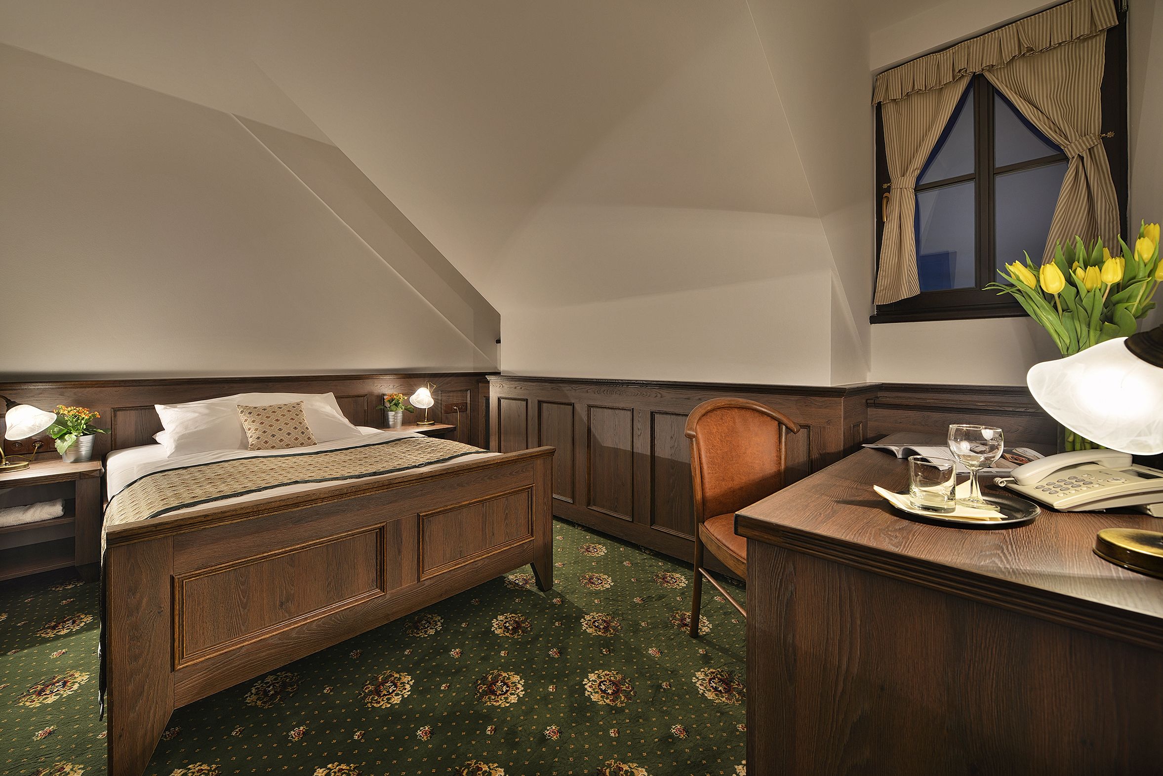 A bedroom with a wooden bed, a desk with a telephone, and a window with curtains.