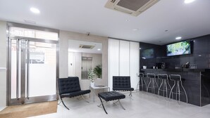 A modern reception area with a bar, two black chairs, a small table, and a TV screen. - Di Carlo Hotels Madrid (Madrid)