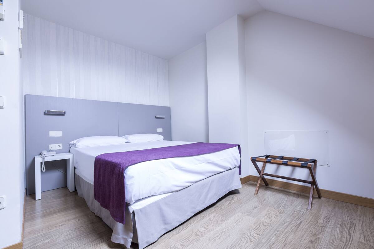 Une chambre bien rangée, avec un lit, une table de chevet et un petit tabouret en bois.