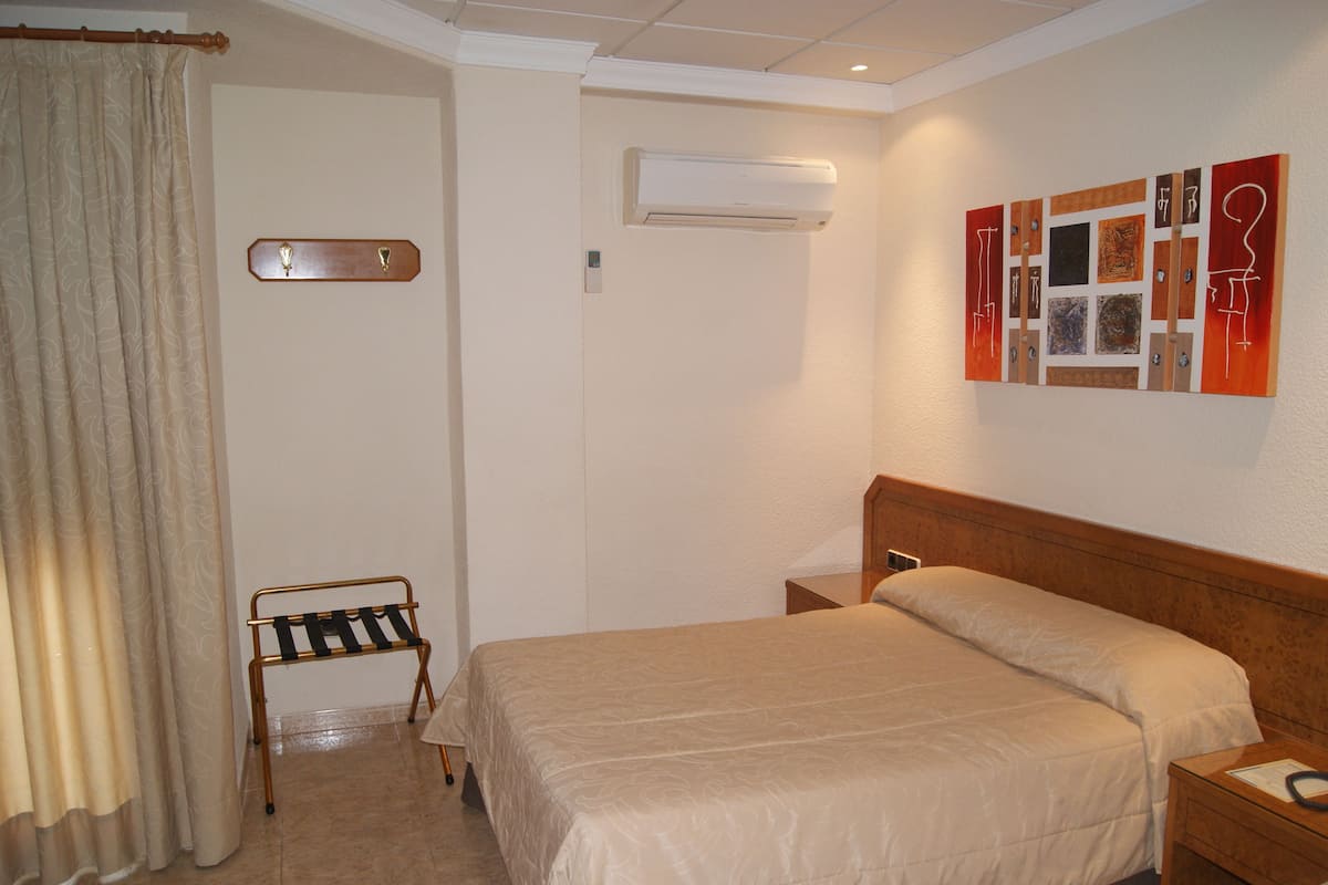 Una habitación de hotel con cama, mesita de noche, aire acondicionado de pared, perchero y cortina.