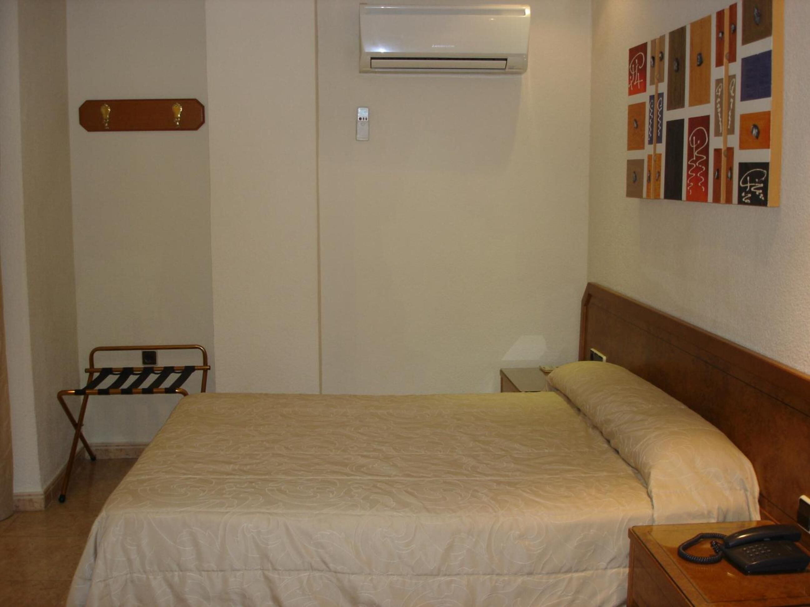 Une chambre d’hôtel comprenant un lit, un appareil de climatisation, une table de chevet en bois avec un téléphone et une œuvre d’art accrochée au mur.