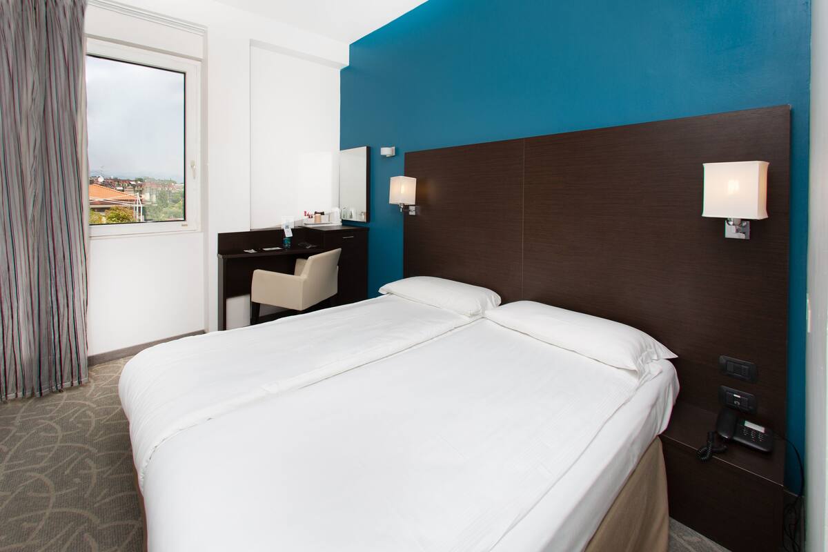 Une chambre d’hôtel avec un grand lit, un bureau et une chaise. Il y a une fenêtre avec des rideaux, une lampe fixée au mur et un téléphone mural.