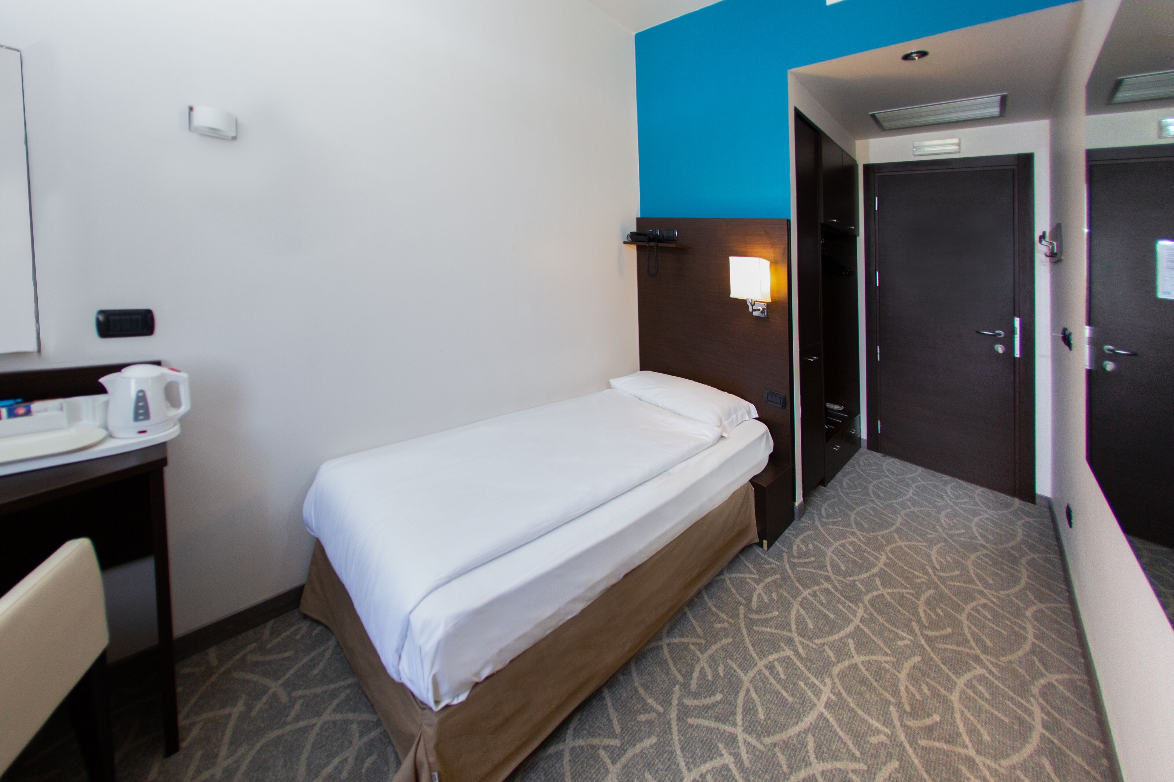 Une chambre d’hôtel avec un lit simple, un bureau avec une machine à café, une lampe fixée au mur et une porte.