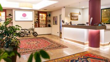 A hotel lobby with a reception desk, a motorcycle, a large rug, and a potted plant.
