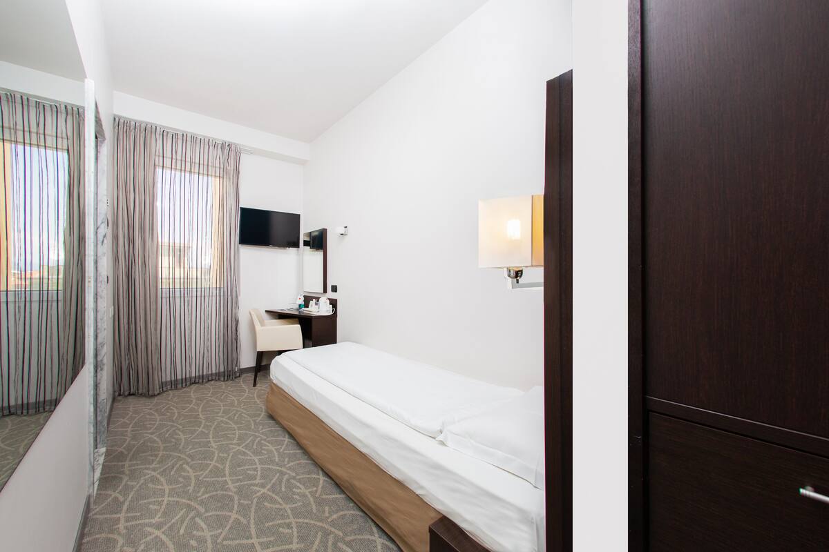 Une chambre d’hôtel avec un lit simple, un bureau avec une chaise, un téléviseur à écran plat fixé au mur et une fenêtre avec des rideaux.