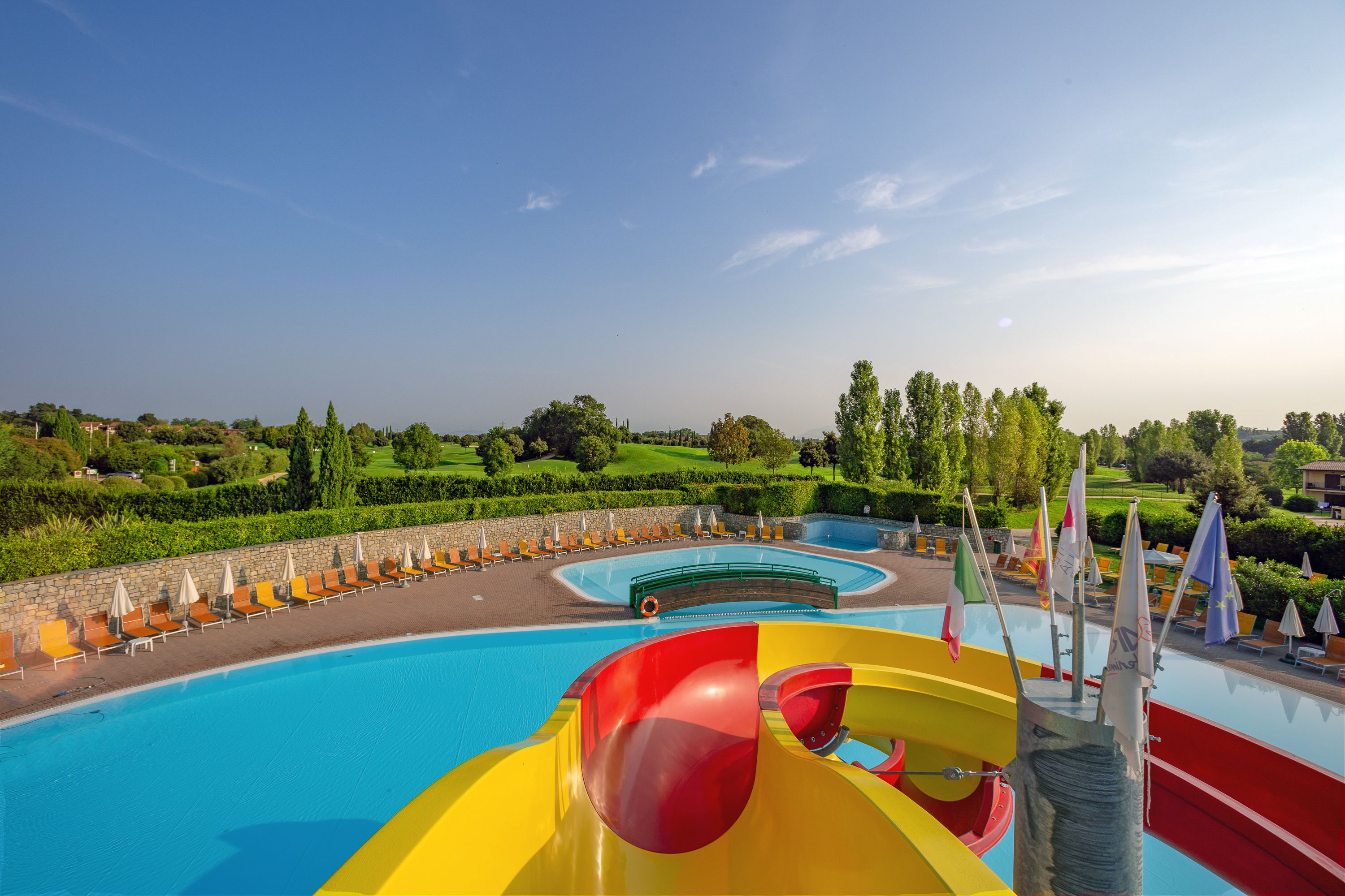 Una piscina con tobogán de agua, sillas de descanso y banderas.