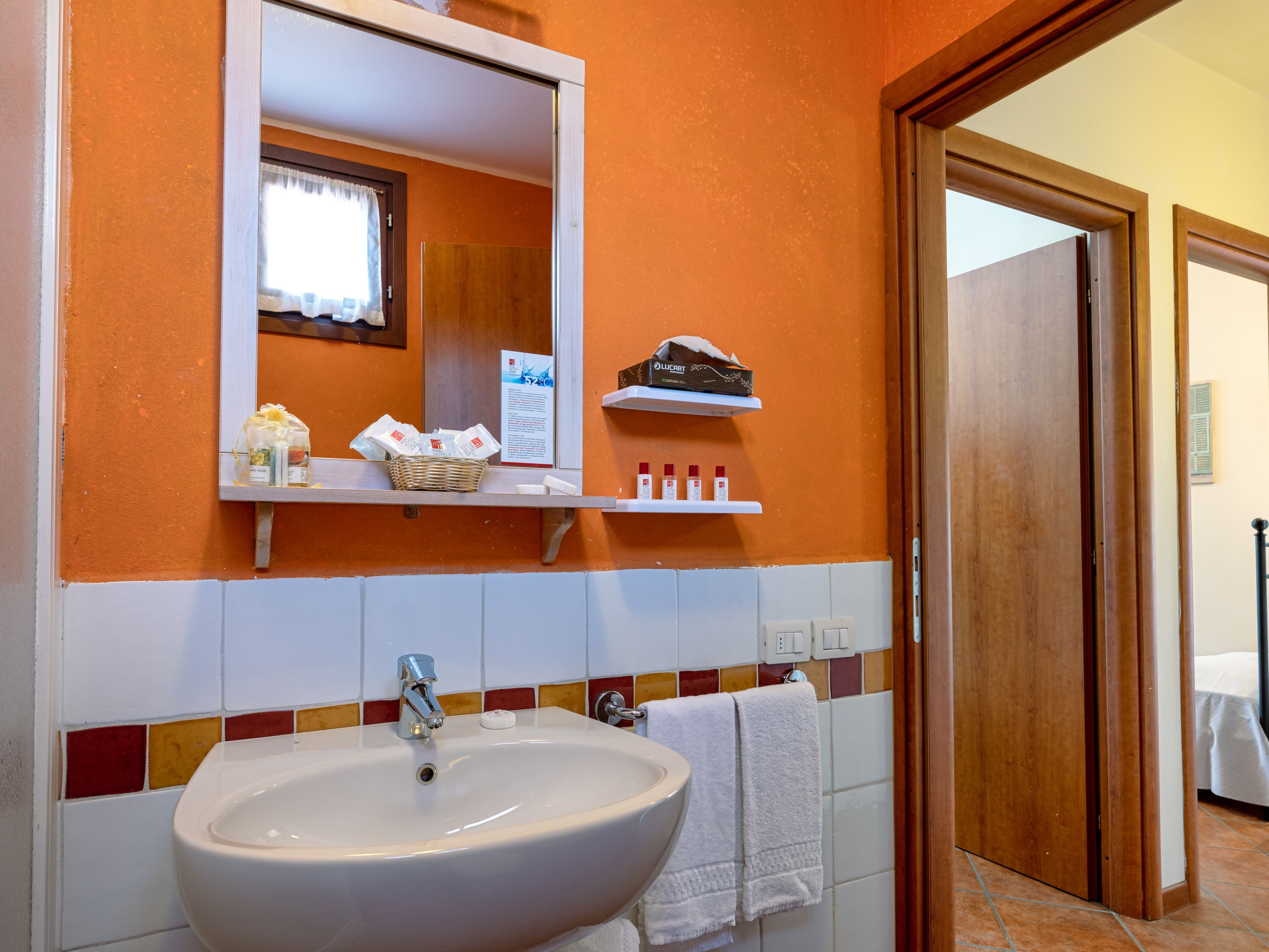 Een badkamer met een wit wastafel, een spiegel en oranje wanden. Er staan toiletartikelen op een plank en een handdoek hangt aan de muur.