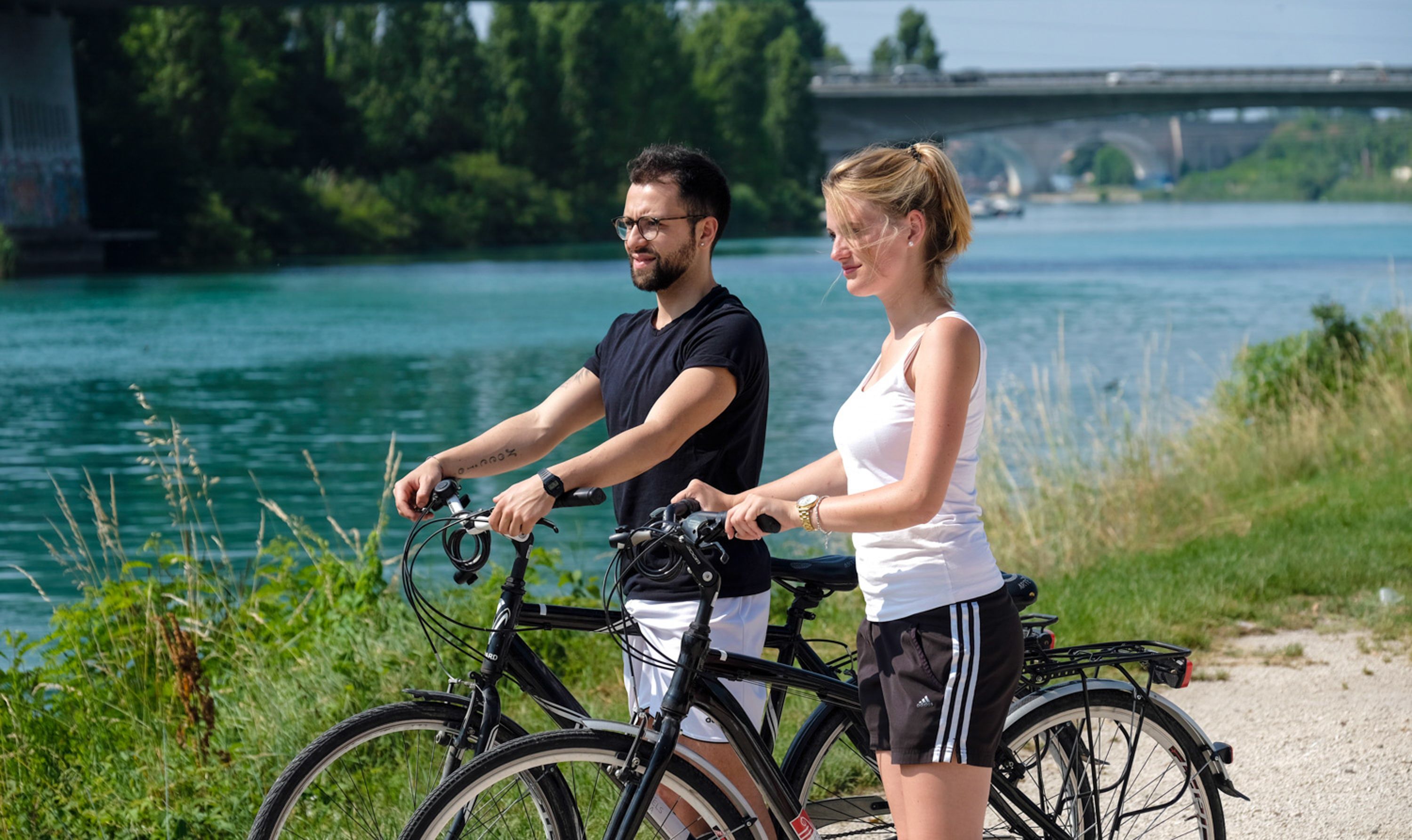Twee personen op fietsen langs een rivier, met een brug op de achtergrond.