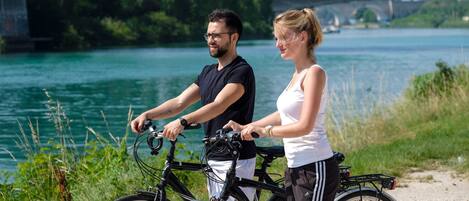 Twee personen op fietsen langs een rivier, met een brug op de achtergrond.