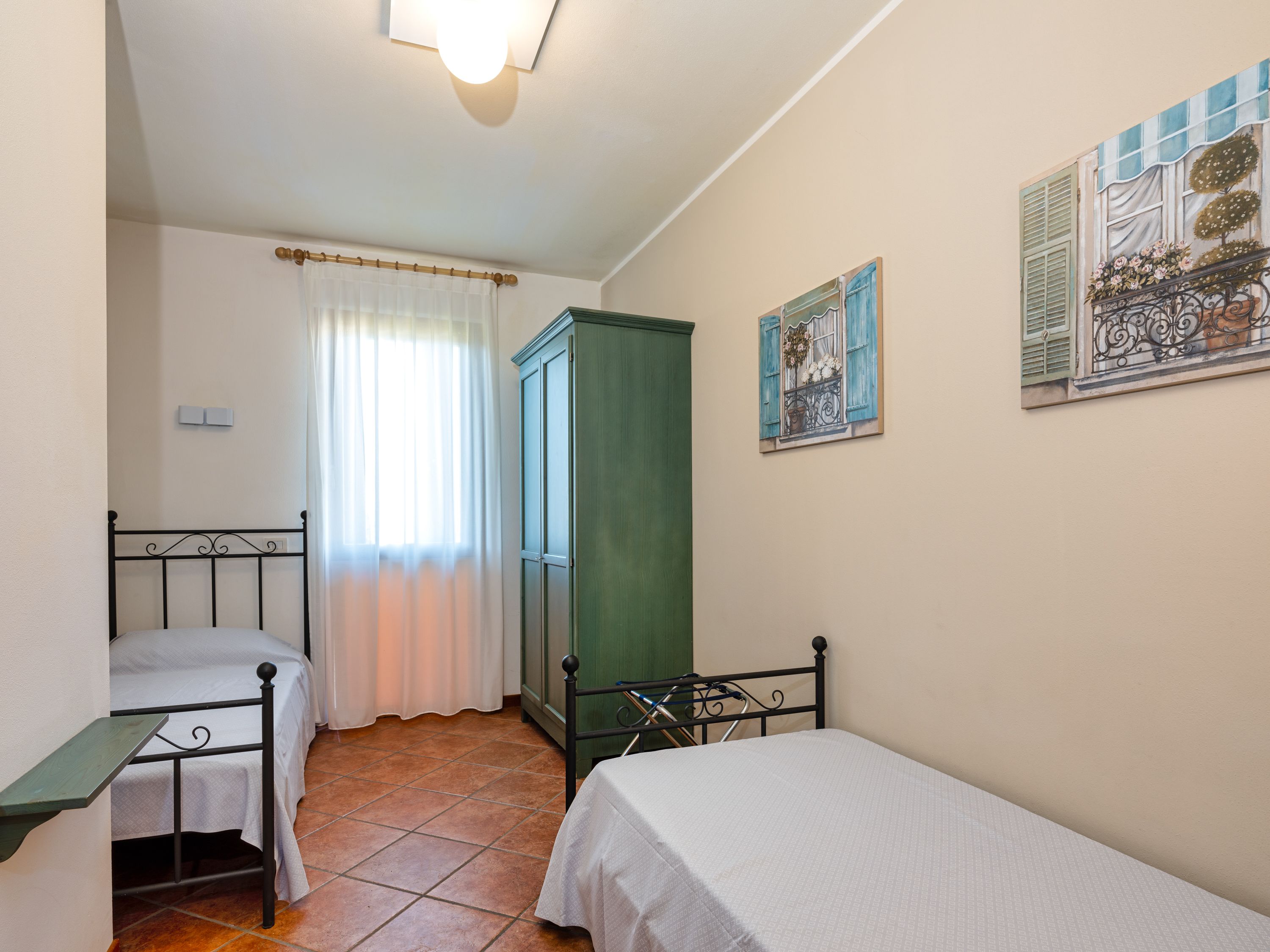 A bedroom with two single beds, a green wardrobe, a window with white curtains, and a wall-mounted painting.