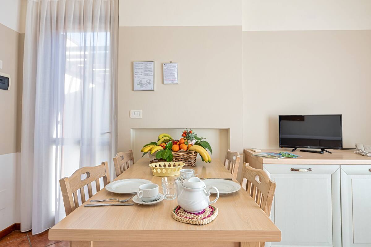 A dining area with a wooden table set for two, a fruit basket, a white pitcher, and a television in the background.