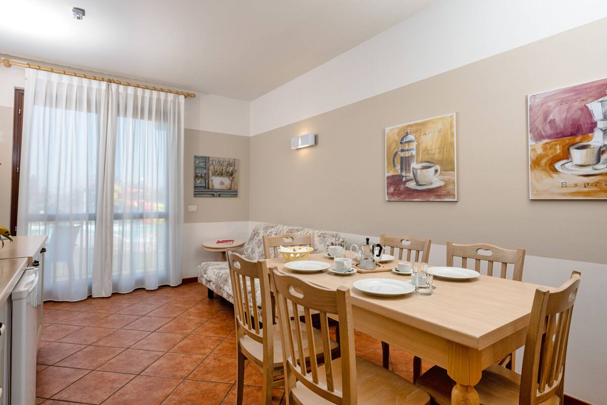 A dining area with a wooden table set for four, a sofa, and a window with sheer curtains.