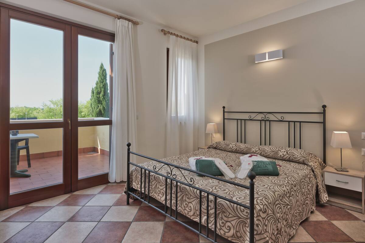 A bedroom with a bed, two bedside tables, a window with curtains, and a view of trees.