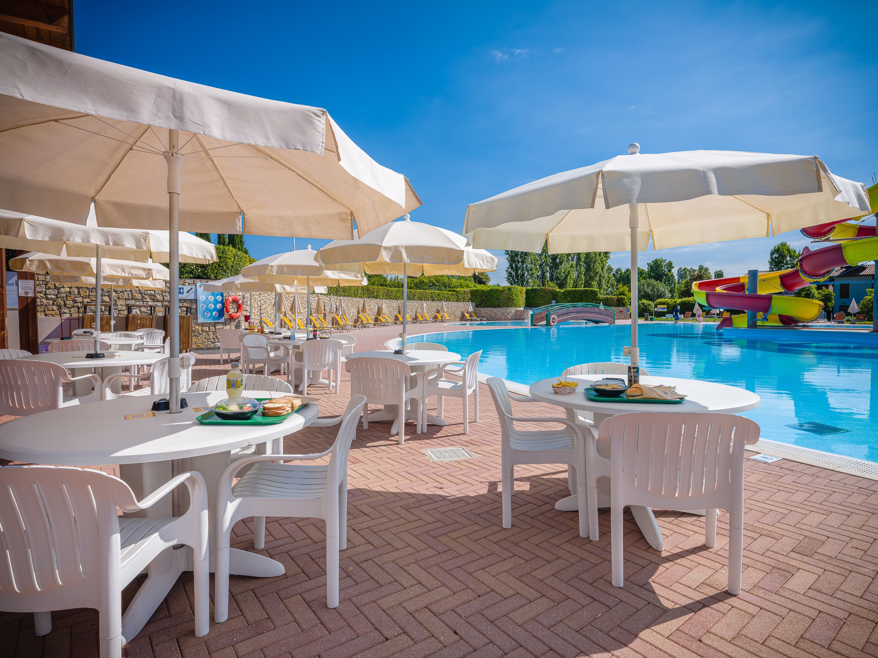 Un espace aménagé au bord de la piscine, avec des chaises et des tables en plastique blanc, des parasols et un toboggan aquatique en arrière-plan.