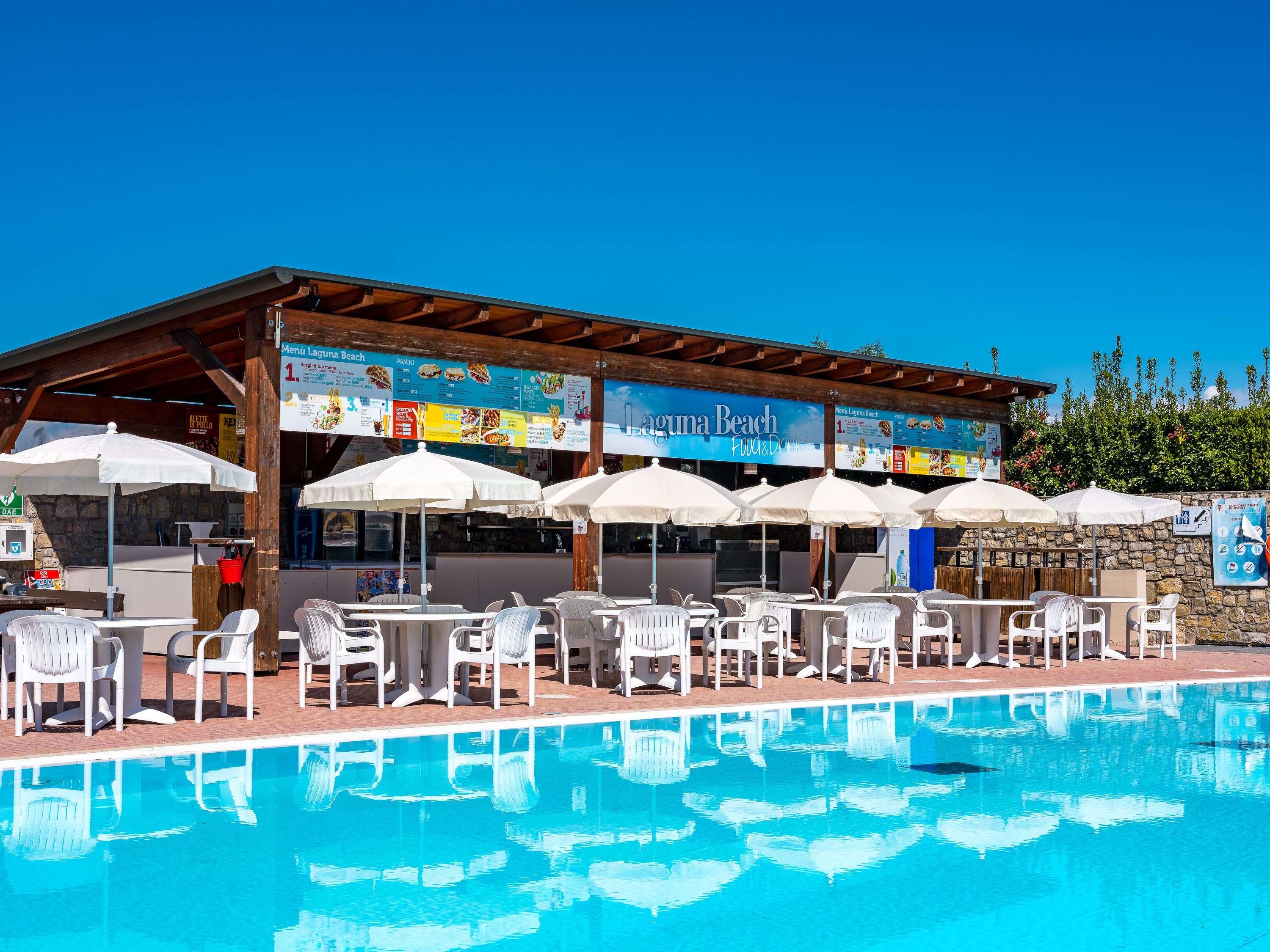 A poolside area with white chairs and umbrellas, a covered seating area with a menu board, and a stone wall in the background.