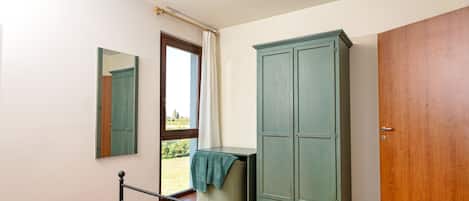 A bedroom with a bed, a wardrobe, a mirror, and a window with a view of greenery.