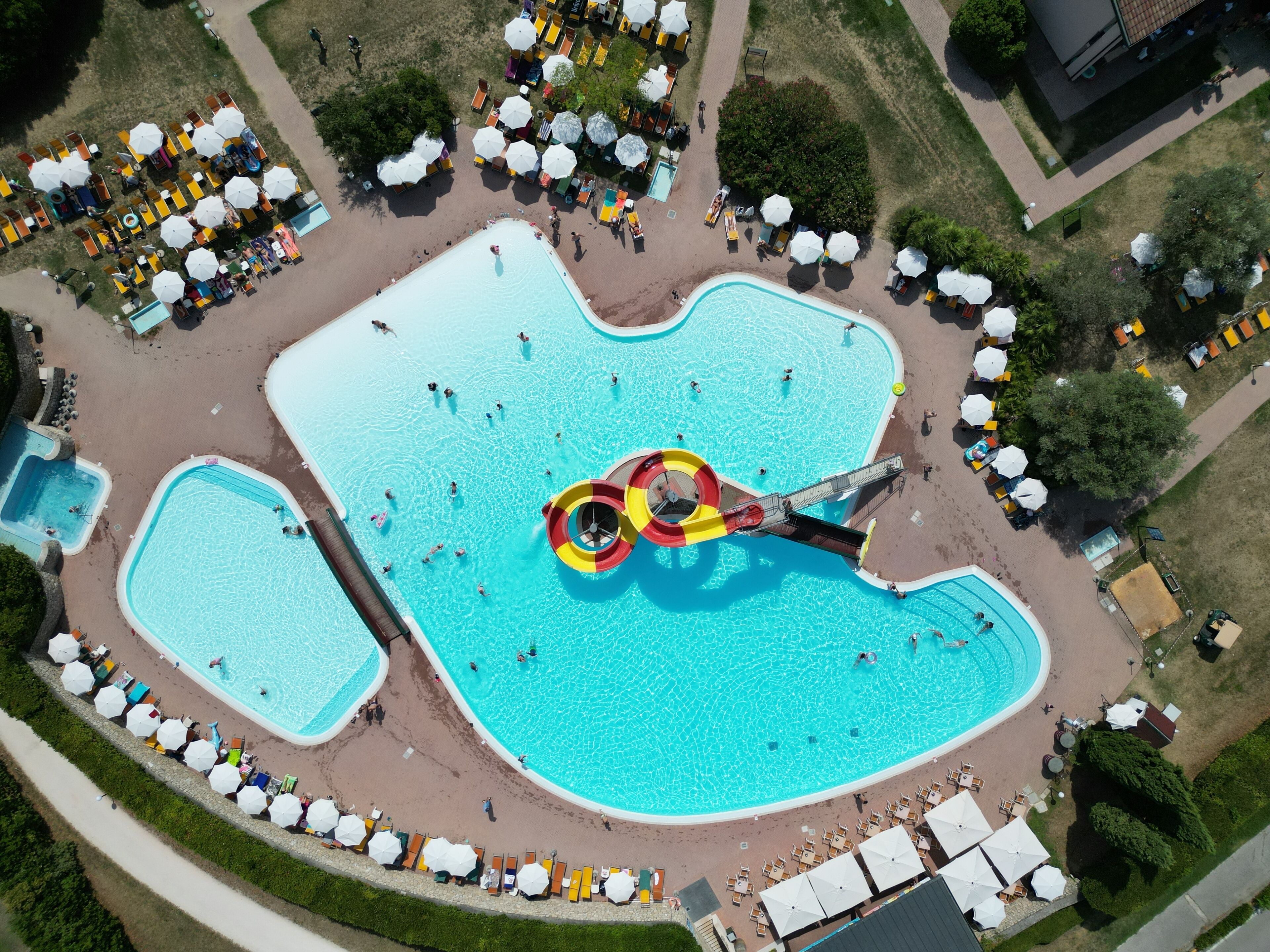 Vue aérienne d’un complexe de piscines comprenant plusieurs bassins, un toboggan aquatique et des espaces de détente aménagés autour.