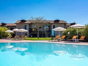 A swimming pool with lounge chairs and umbrellas, a building with a tiled roof, and a clear blue sky. - Residence Eden (Castelnuovo del Garda)
