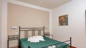 Un lit simple avec une couvre-lit verte, un cadre en métal noir et des oreillers blancs. Il y a une petite table de chevet avec une lampe et un interrupteur fixé au mur. Un vase décoratif est accroché au mur au-dessus du lit.