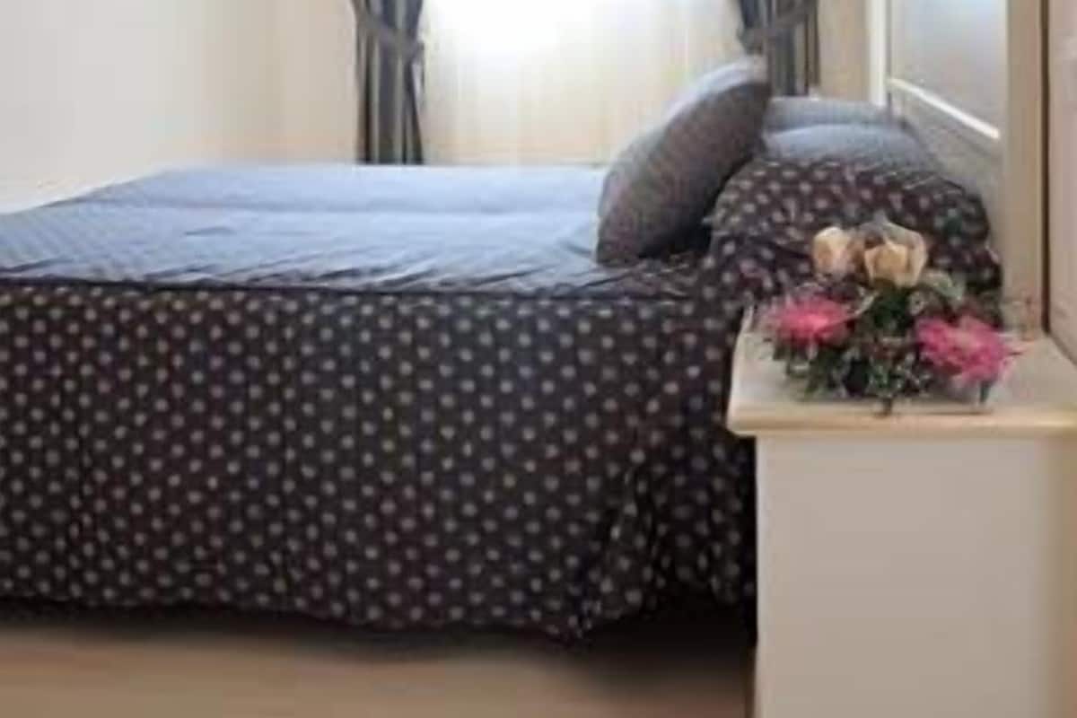 Ein Schlafzimmer mit Bett, Vorhängen, einem Nachttisch mit Blumenvase und einem Spiegel.