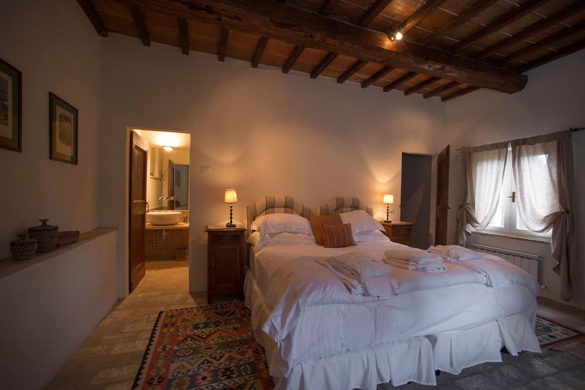 A bedroom with a large bed, two bedside tables with lamps, a wooden ceiling, a bathroom visible through an open door, and a patterned rug.
