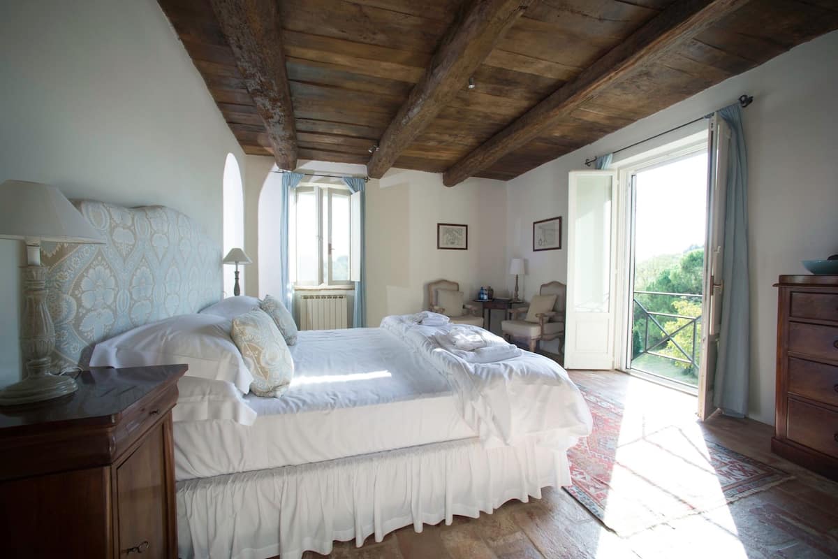 A bedroom with a large bed, wooden ceiling beams, a dresser, and a view of greenery through the open door.