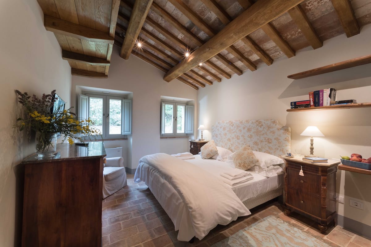 A bedroom with a wooden ceiling, a bed with white bedding, a nightstand with a lamp, and a bookshelf.