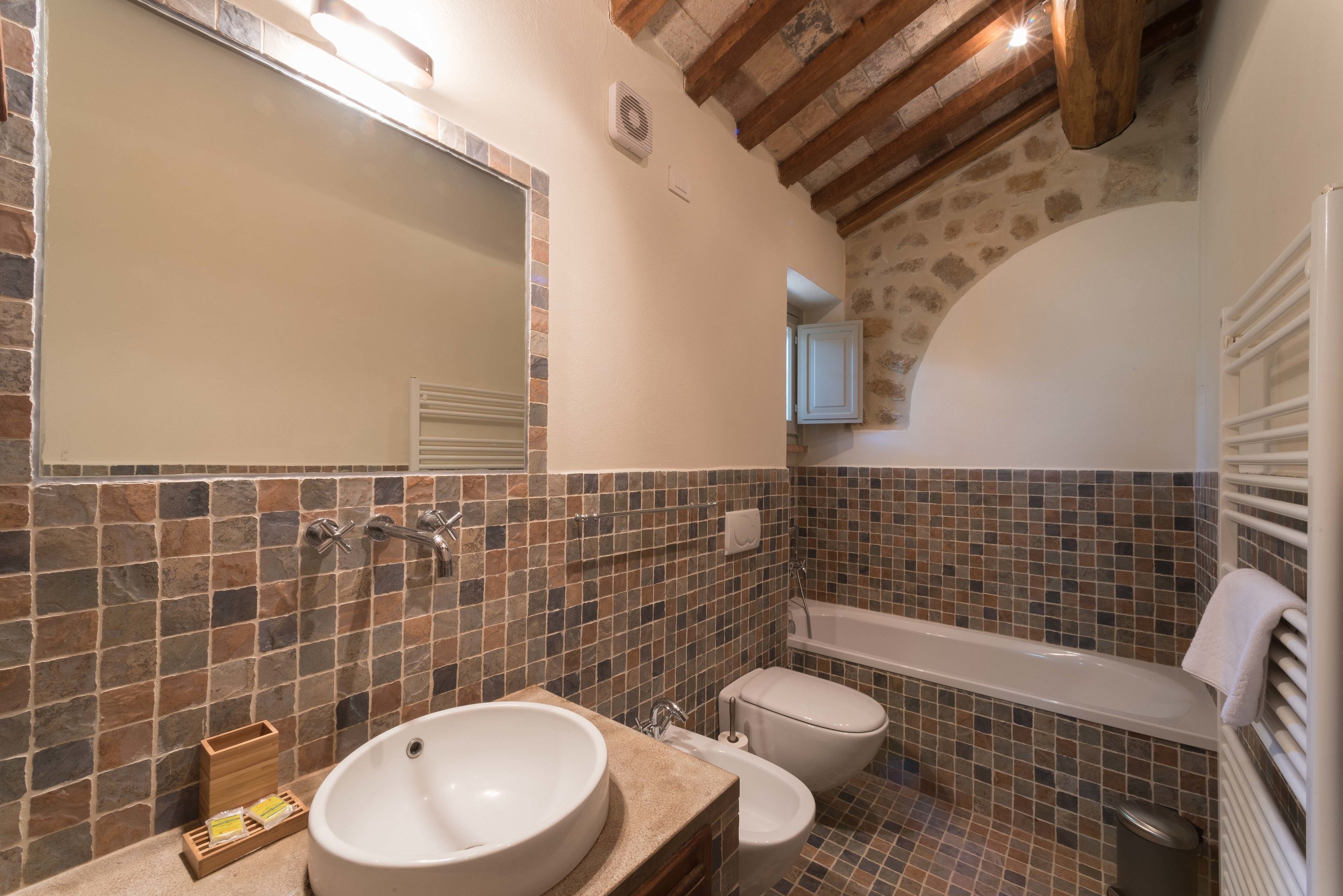 Un baño con lavamanos grande, inodoro y bañera, con paredes de mosaico y vigas de madera.